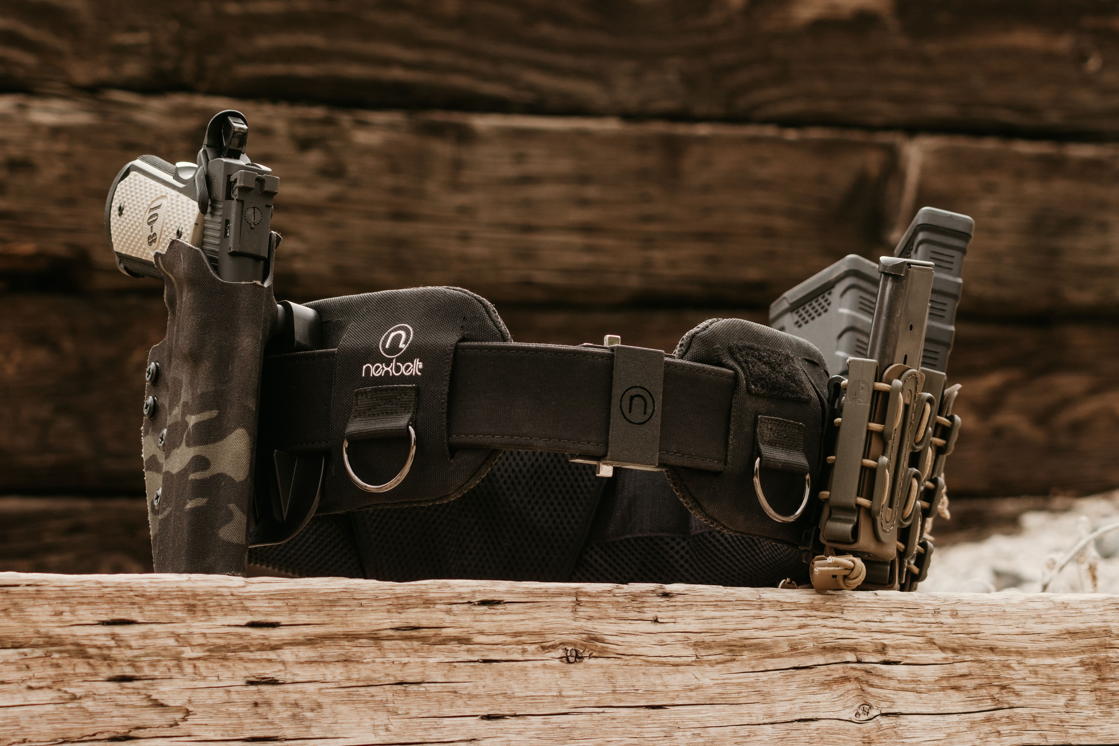A black tactical rig battle belt sits against a wood background, the belt has several artillery pieces clipped to it, as well as the Nexbelt logo - battle belt vs chest rig | tactical rig belt